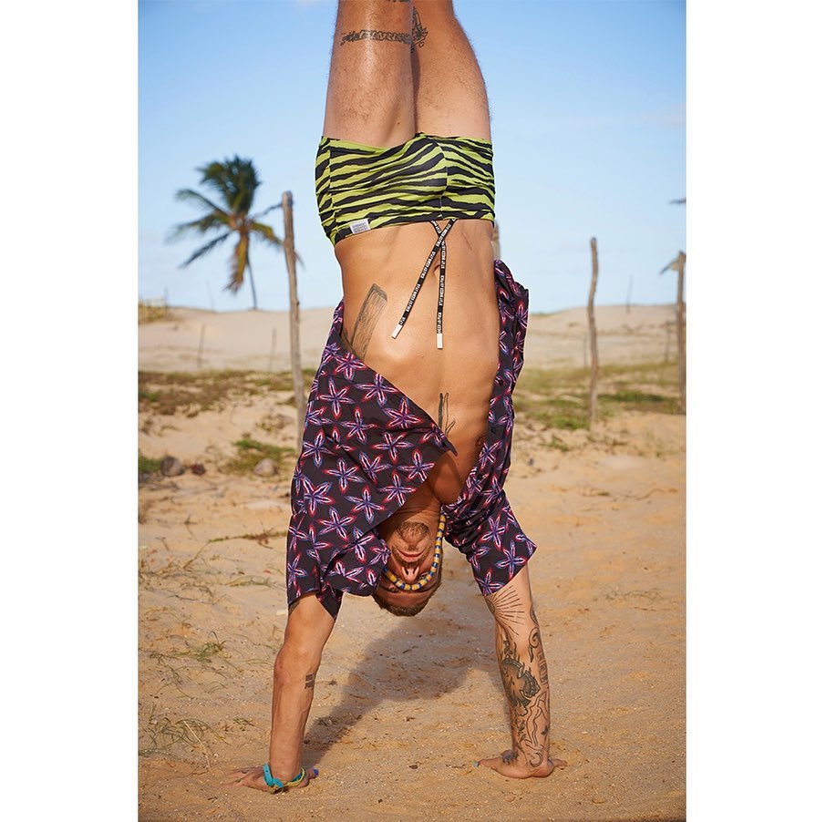 Person doing a handstand on a beach wearing a patterned shirt and green and black patterned shorts near a palm tree on a sunny day.
