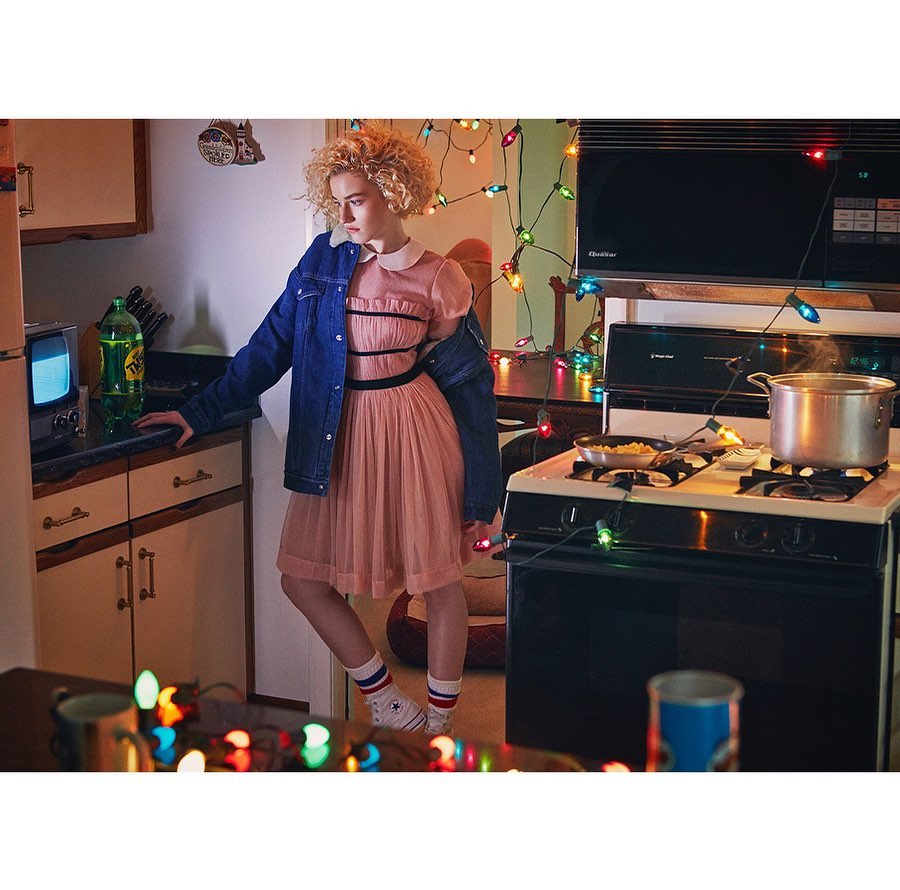 Retro-style portrait of a young woman in a pink dress and denim jacket, standing in a kitchen with vintage appliances and Christmas lights.