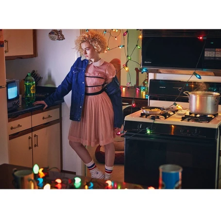 Retro-style portrait of a young woman in a pink dress and denim jacket, standing in a kitchen with vintage appliances and Christmas lights.