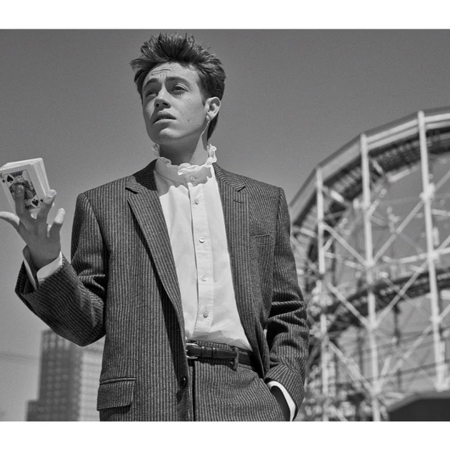 Monochrome portrait of a young man in a pinstripe suit holding a deck of cards with a building structure in the background.