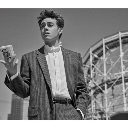 Monochrome portrait of a young man in a pinstripe suit holding a deck of cards with a building structure in the background.