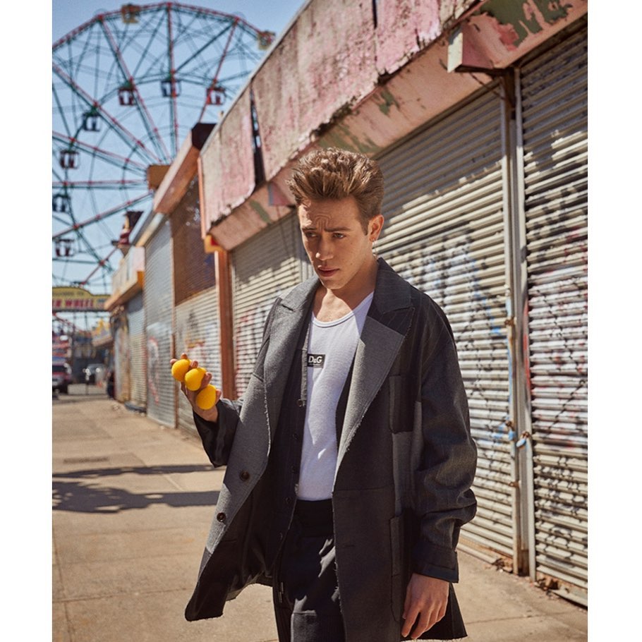 Fashionable young man with lemons stands on a weathered street near a colorful Ferris wheel, wearing a grey coat and white graphic t-shirt.