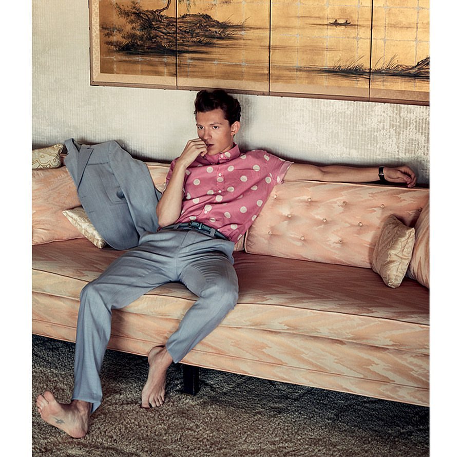 Adult male rests on a light pink sofa wearing pink polka dot short-sleeved shirt, light blue pants, and barefoot in a light-filled room.