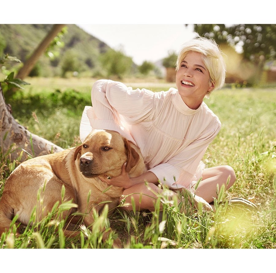 Woman in a light pink dress smiles while sitting in a grassy field hugging a yellow labrador retriever dog in natural outdoor light.