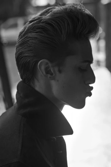 Monochrome portrait of a young adult male with stylized hair, wearing a dark coat, posing in profile against an out-of-focus background.