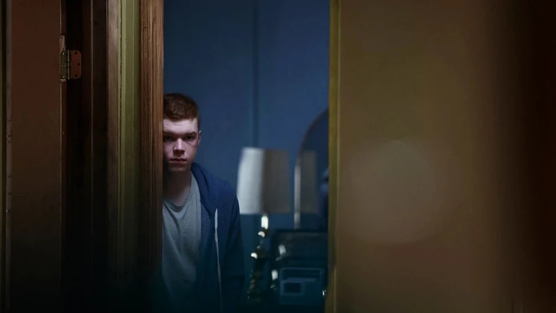 A young adult man peeks out from behind a wooden door, looking serious with a blue hoodie, in an indoor setting with muted lighting.