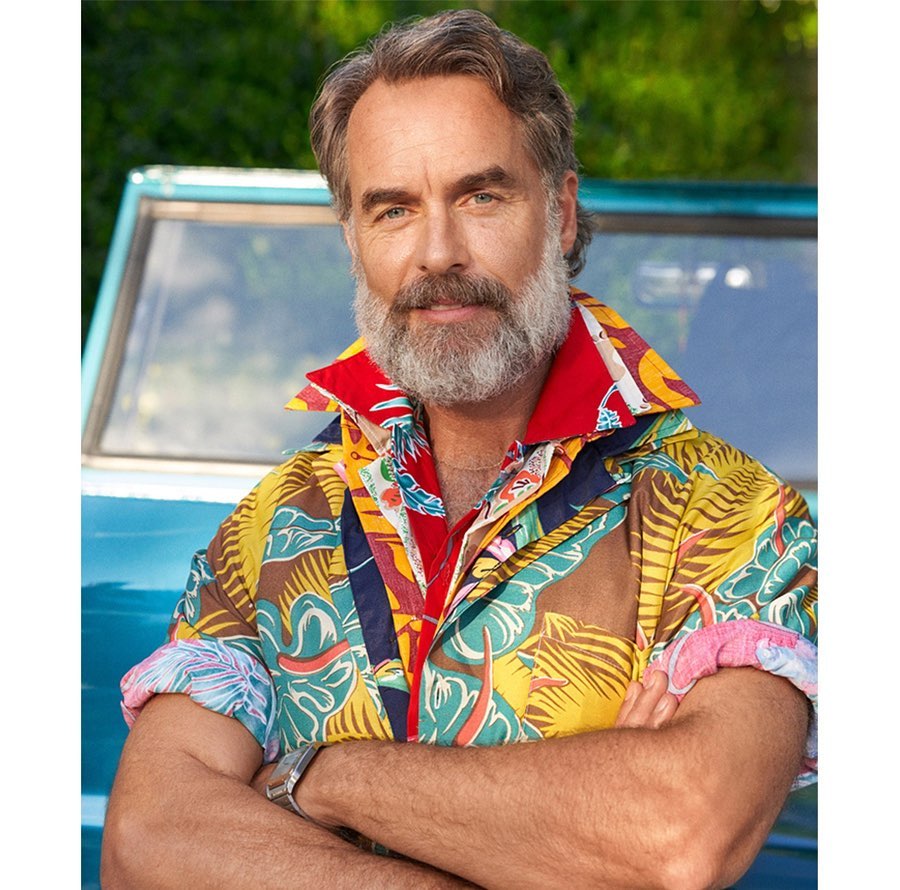 Portrait of a smiling mature man with a grey beard, wearing a brightly colored patterned shirt, standing outdoors in front of a blue vehicle.