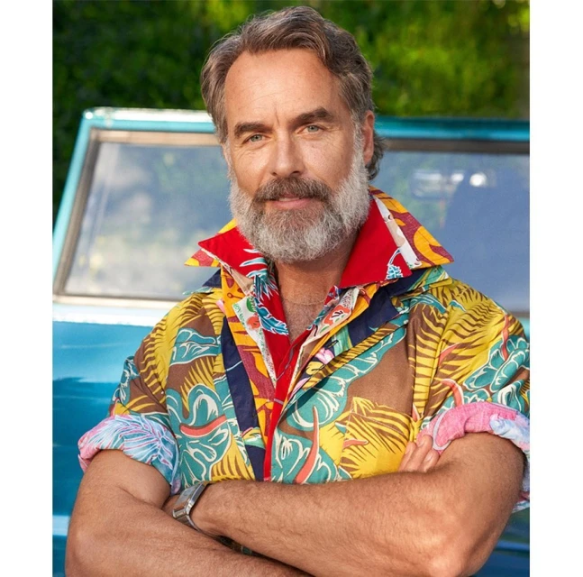 Portrait of a smiling mature man with a grey beard, wearing a brightly colored patterned shirt, standing outdoors in front of a blue vehicle.