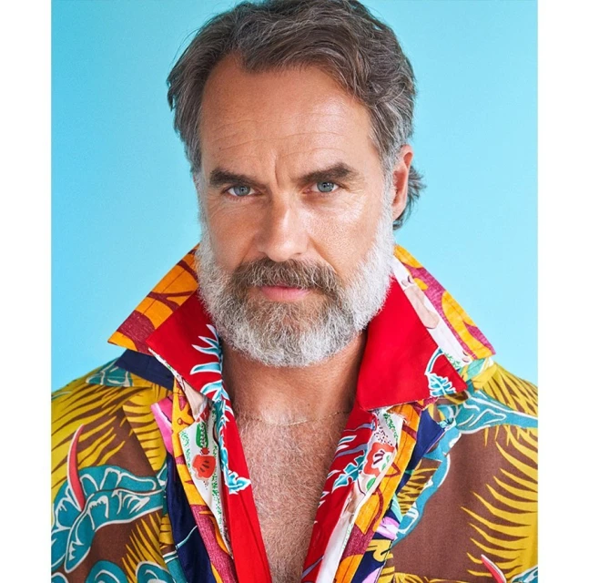 Portrait of a mature adult male with gray hair and beard wearing a brightly patterned short-sleeved shirt against a light blue backdrop.