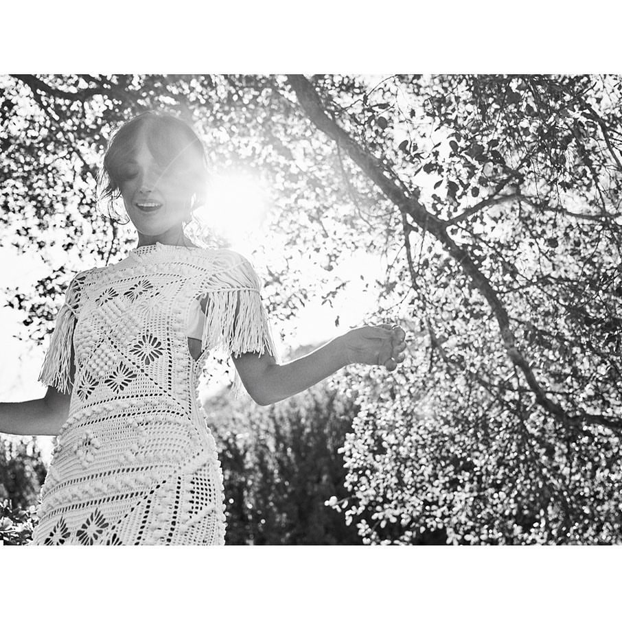 Black and white photo of a woman in a dress with fringe standing outside beneath a tree with a bright light in the background.