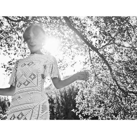 Black and white photo of a woman in a dress with fringe standing outside beneath a tree with a bright light in the background.
