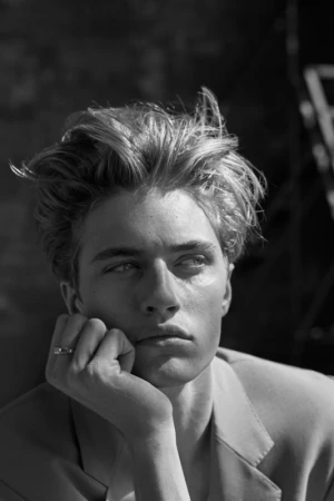 Black and white portrait of a blonde man resting his chin on his hand. He wears a blazer and ring, with a blurred outdoor background.