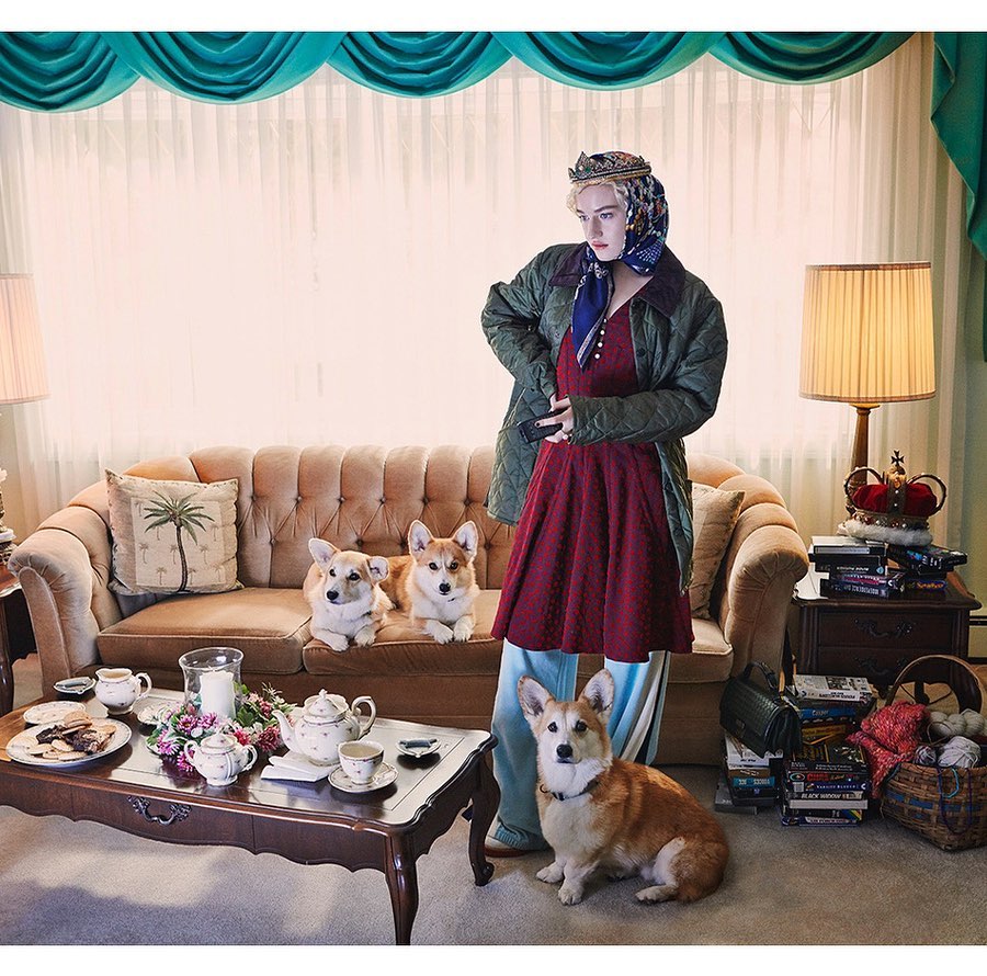 Woman dressed as Queen Elizabeth stands with Corgi dogs in a traditionally decorated living room with a couch and tea service.