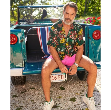 Bearded man sits on the back of a teal convertible car wearing a floral shirt and pink shorts, surrounded by greenery and sunlight.