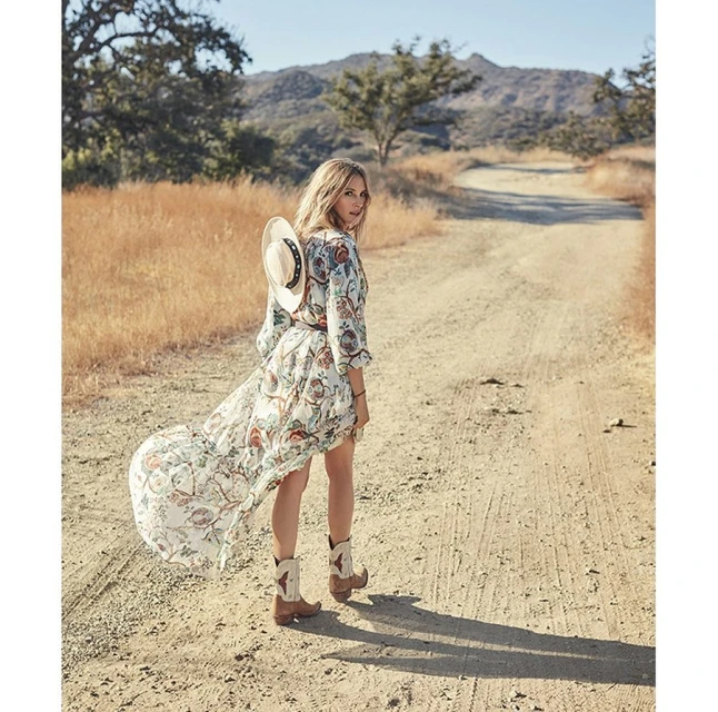A woman in a floral dress and cowboy boots walking on a dirt road with a cowboy hat slung over her shoulder in a rural setting.