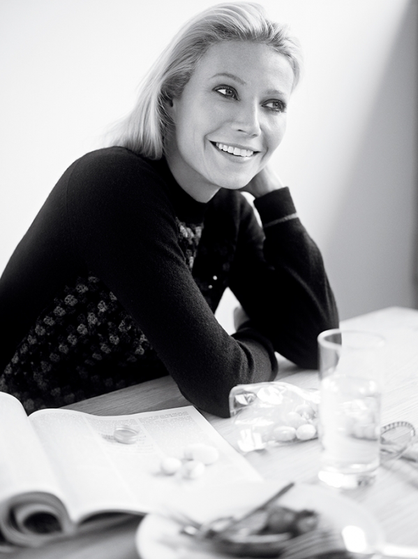 Black and white portrait of Gwyneth Paltrow smiling while seated at a wooden table with an open magazine