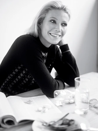 Black and white portrait of Gwyneth Paltrow smiling while seated at a wooden table with an open magazine