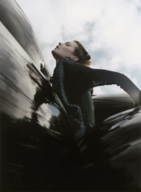 Low-angle shot of a woman in a dark top with her head tilted back against a shiny black surface, with the sky in the background.