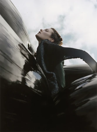 Low-angle shot of a woman in a dark top with her head tilted back against a shiny black surface, with the sky in the background.