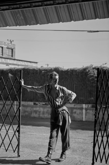 Black and white portrait of a young man in an outdoor setting, wearing a striped shirt and standing near a metal gate.