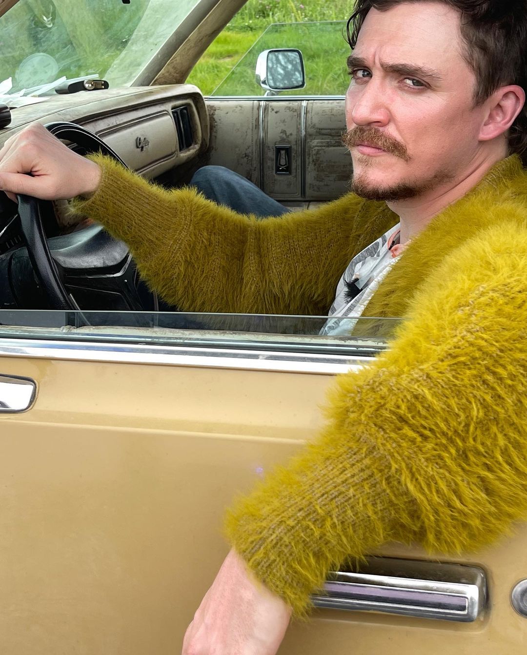 Man in yellow sweater sits in a vintage car. Driver holds steering wheel while looking at the camera in daylight with greenery visible outside.