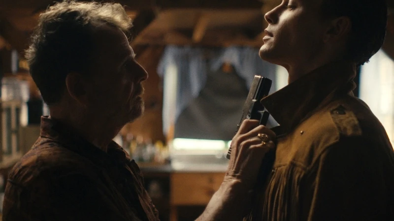 An intense moment unfolds indoors: a mature man holds a gun to a younger man's neck, both figures partially silhouetted against a blurred backdrop.