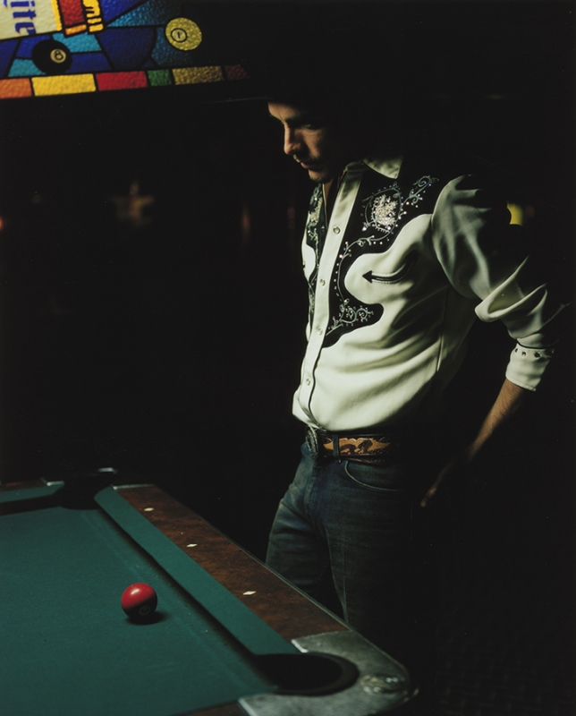 Man stands near a billiards table in a dimly lit room, wearing a western-style shirt and jeans, next to a green pool table with a red ball.