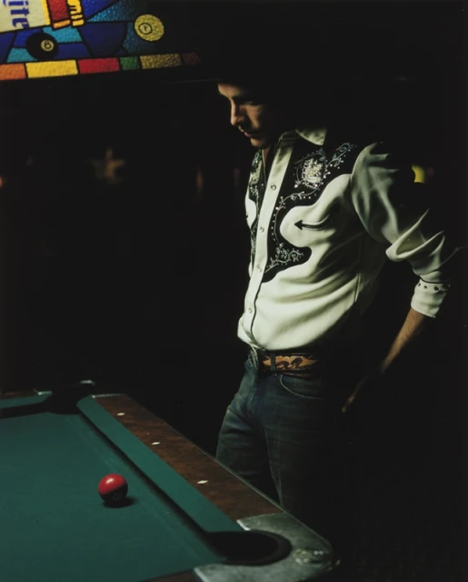 Man stands near a billiards table in a dimly lit room, wearing a western-style shirt and jeans, next to a green pool table with a red ball.