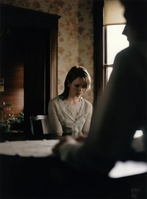 Woman sits at a table with papers. A man's silhouette is in the foreground. The setting is indoors, with floral wallpaper.