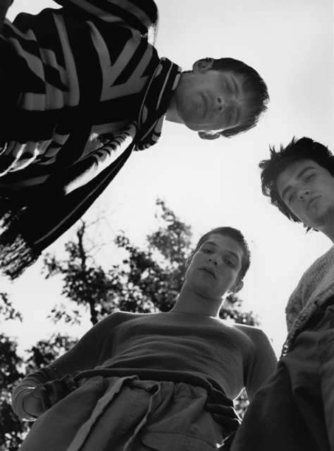Low-angle, black and white shot of three young adult males looking down at the camera, with trees and bright sky in the background.