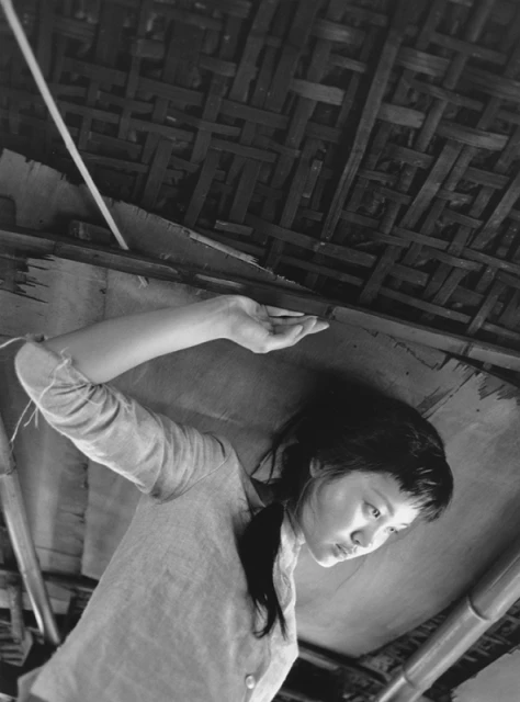 Monochrome portrait of a woman looking down, with her arm raised and a woven ceiling in the background, creating a thoughtful mood.