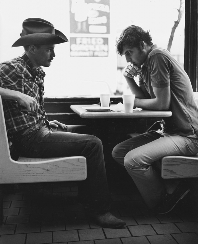 Two men sit at a table indoors; one in a cowboy hat, in black and white, creating a vintage and intimate atmosphere.