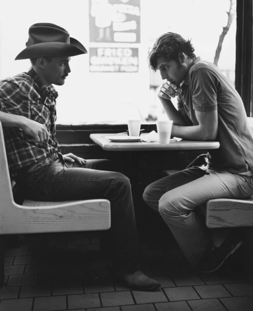 Two men sit at a table indoors; one in a cowboy hat, in black and white, creating a vintage and intimate atmosphere.