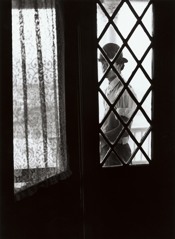 Monochrome portrait of a person in hat behind diamond pane window, with lace curtain visible nearby, creating an intimate indoor scene.