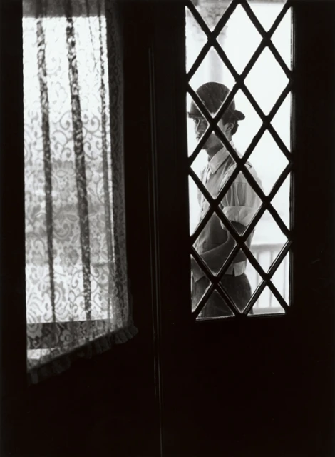 Monochrome portrait of a person in hat behind diamond pane window, with lace curtain visible nearby, creating an intimate indoor scene.