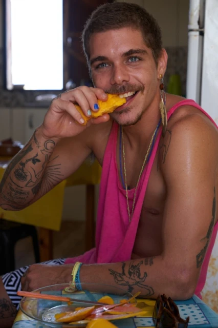 A young adult man with tattoos, wearing a pink tank top, happily eats a piece of mango indoors, showcasing painted fingernails and casual style.