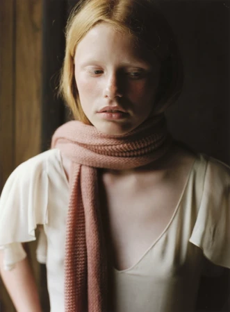 Close-up portrait of a fair-haired young woman wearing a pale pink knit scarf and an off-white top against a dark interior background.