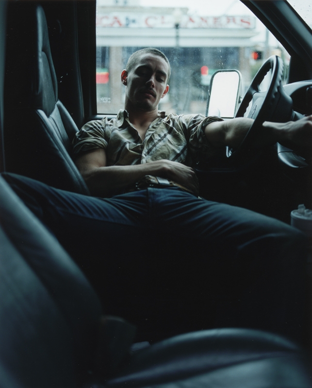 A man sits inside a vehicle with his arm resting on the steering wheel, with a building visible through the windshield.