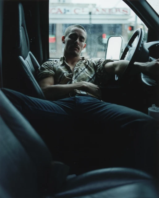 A man sits inside a vehicle with his arm resting on the steering wheel, with a building visible through the windshield.