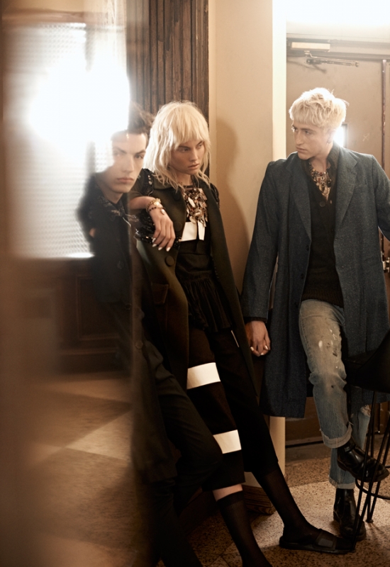 Fashion editorial shot of two models with blonde hair in a dimly lit indoor setting. One leans against a wall while the other stands nearby.