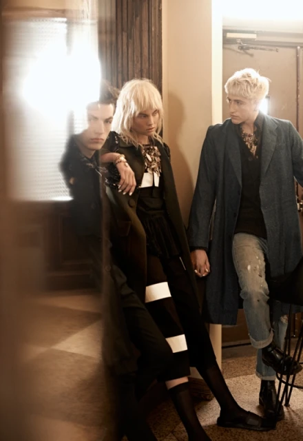 Fashion editorial shot of two models with blonde hair in a dimly lit indoor setting. One leans against a wall while the other stands nearby.