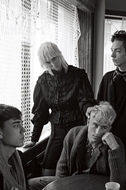 Black and white portrait of four people indoors. Three young men and one woman pose in dark clothing near a window in an editorial style.