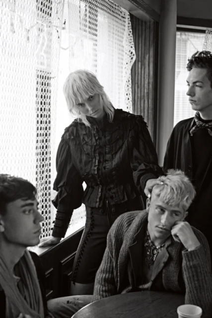 Black and white portrait of four people indoors. Three young men and one woman pose in dark clothing near a window in an editorial style.