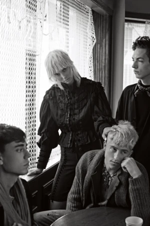 Black and white portrait of four people indoors. Three young men and one woman pose in dark clothing near a window in an editorial style.