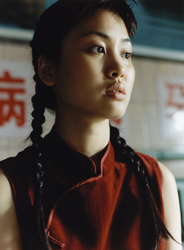 Portrait of a young woman with braided hair wearing a rust-colored sleeveless top, looking off to the side in an indoor setting.
