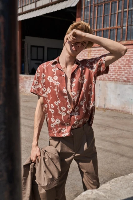 Male fashion model in floral shirt poses in front of industrial building. Outdoor shot with neutral color palette and natural light.