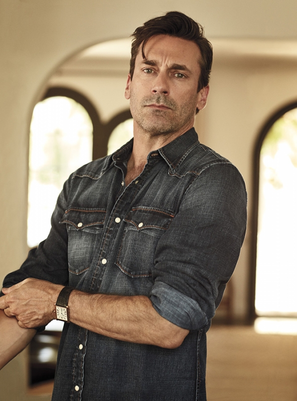Portrait of an adult male with dark hair in a denim shirt with a watch, posing in an indoor environment with an arched doorway.