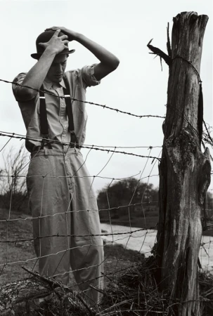 Black and white portrait of a slender adult in a bowler hat, suspenders, and high-waisted pants next to a barbed wire fence.