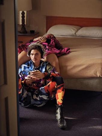 Man in patterned camo outfit seated on floor holding phone with woman resting on bed behind him by Carter Smith