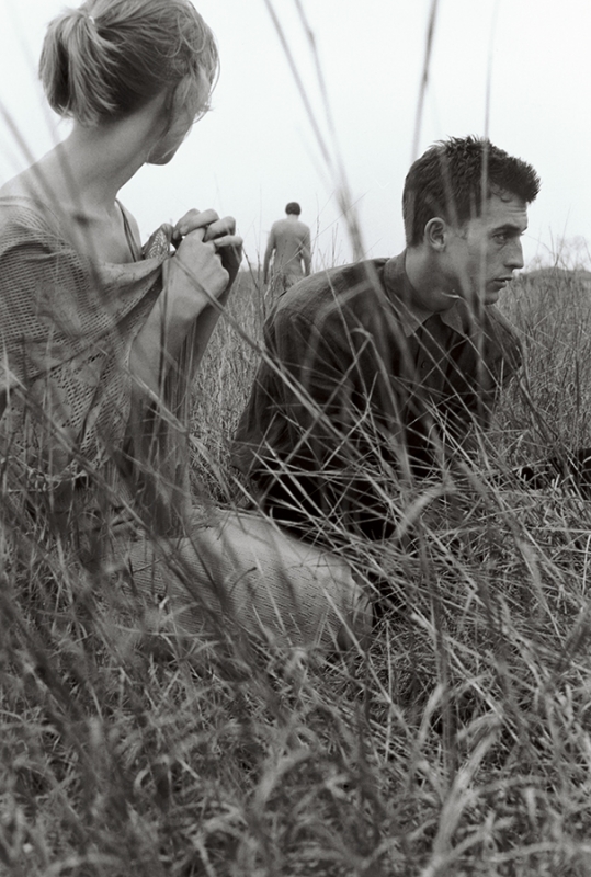 Monochrome portrait of a man and woman in a field of tall grass, with a third person walking in the distance.